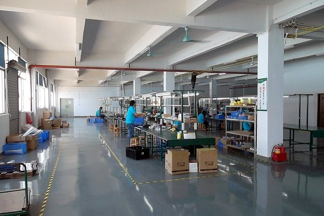 Assembly line with workers along long benches, material racks and cartons alongs…