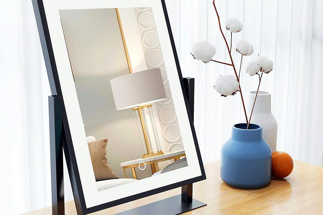 A black-framed rectangular table mirror on a wooden table, reflecting a table lamp, with a blue vase and cotton stems beside it